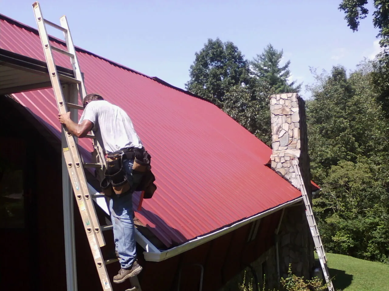 Roofer on a ladder beside a red metal roof and stone chimney — Soffit & Fascia Repair by Roofing Brothers in Vidalia